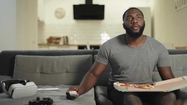 Afrikaner sitzt auf Sofa in offener Küche. Schwarzer Mann isst Pizza. — Stockvideo