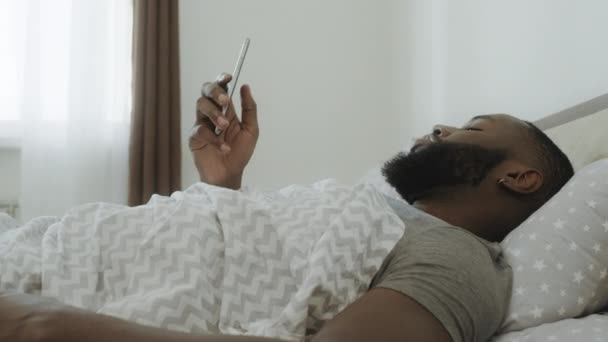 Shocked black man sitting in bed. Smartphone falling out of man hand. — Stock Video