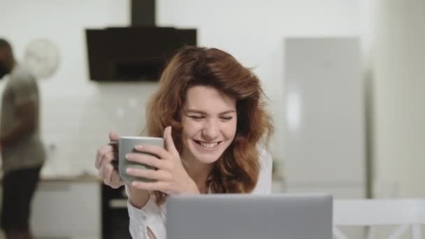 Lächelnde weiße Frau liest freudige Nachrichten am Laptop in der Morgenküche. — Stockvideo