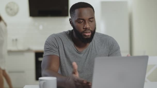Trauriger schwarzer Mann arbeitet Laptop in offener Küche. Verrückter junger Kerl tippt Notizbuch — Stockvideo