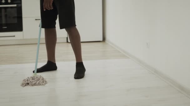 Чернокожий мужчина моет пол на кухне шваброй. Серьезный парень моет пол в носках — стоковое видео