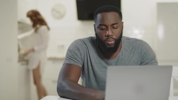 Σοβαρός μαύρος άνθρωπος που εργάζεται στο φορητό υπολογιστή στην ανοιχτή κουζίνα. Χαμογελαστός ζευγάρι που πίνει τσάι — Αρχείο Βίντεο