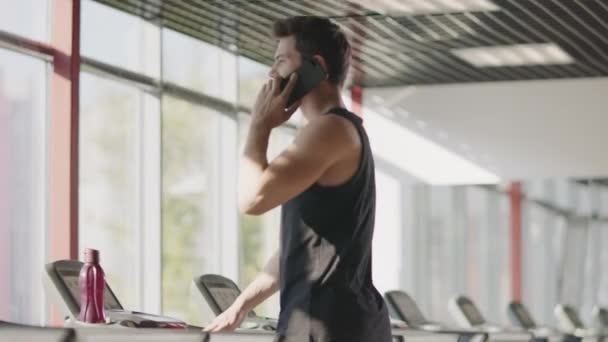 Pohledný muž používající mobilní telefon v tělocvičně. Velký sportovec v běžecké trenažéru — Stock video