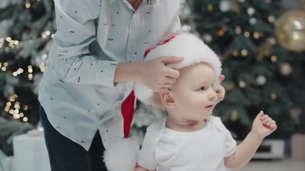 Um rapazinho a pôr o chapéu do Pai Natal na sala de estar. Irmão mais velho usando chapéu xmas — Vídeo de Stock