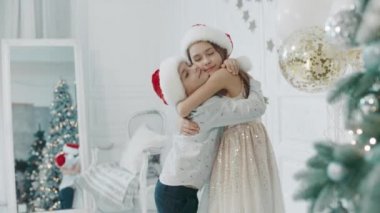 Chritmas ağacının yanında birbirlerine sarılan gülümseyen çocuklar.
