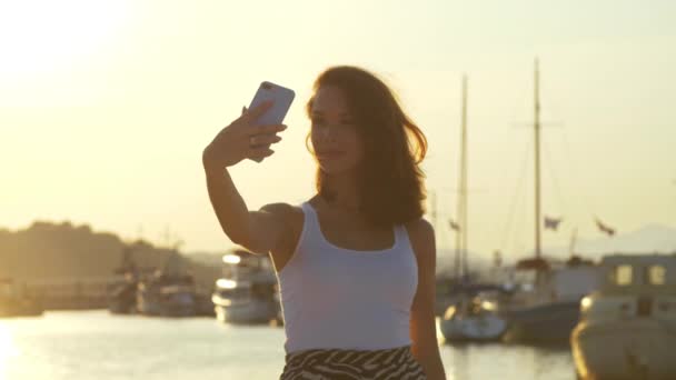 Mujer feliz tomando fotos móviles en el puerto de la tarde. Chica de viaje posando para selfie — Vídeo de stock