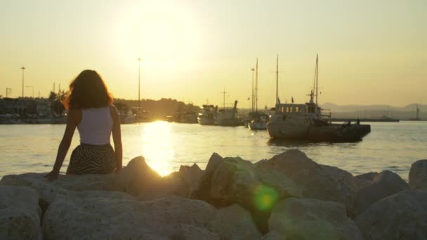 Toeristische meisje op steen bij avond zonsondergang in zeehaven. Vrouw op zonsondergang landschap — Stockvideo