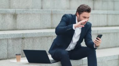 İş adamı sokakta telefon kamerasına el sallıyor. Açık havada sohbet eden bir adam.