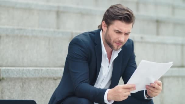 Üzletember vizsgálja az üzleti papírokat a szabadban. Férfi menedzser ül a lépcsőn — Stock videók