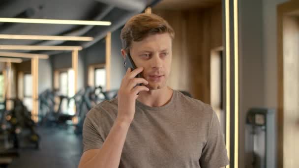 Красивий спортсмен розмовляє по телефону в спортивному клубі. Бодібілдер стоїть в спортзалі . — стокове відео