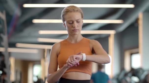 Chica atleta molesta usando smartwatch en el gimnasio. Deportiva de pie en el club deportivo — Vídeos de Stock