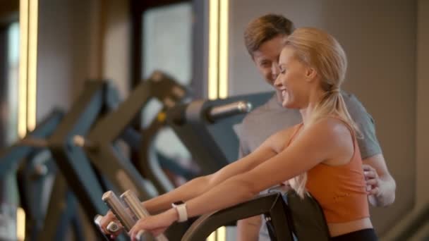 Hot fit meisje maken biceps krullen in sportclub. Fit man helpt zijn cliënt in de sportschool — Stockvideo
