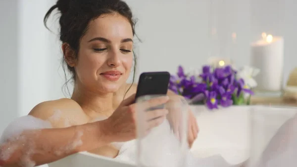 Close up smiling woman chatting on smartphone in bathtub in slow motion.
