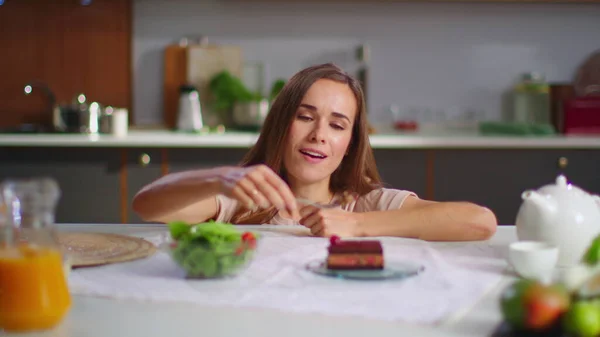 Onnellinen nainen syö kakkua sen sijaan salaattia keittiössä. Lady nauttii kakku pöydässä — kuvapankkivalokuva