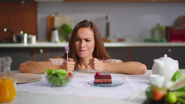 Nainen valitsemalla kakku sijaan salaattia keittiössä. Nainen syö kakkua keittiössä — kuvapankkivalokuva