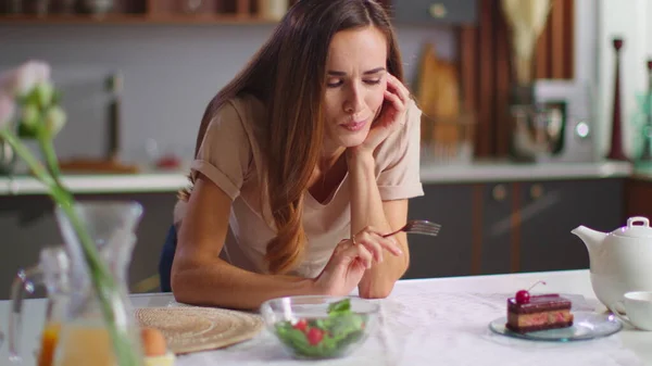 Thoughtful woman choosing between cake and salad on kitchen