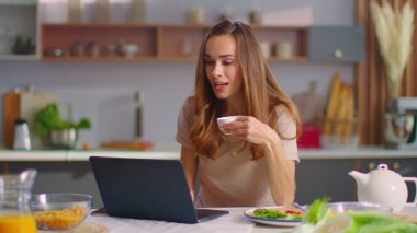 Woman chatting on webcam with friend on laptop at kitchen. Lady drinking coffee