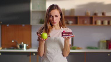 Woman making choice between apple and cake on kitchen. Woman choosing apple