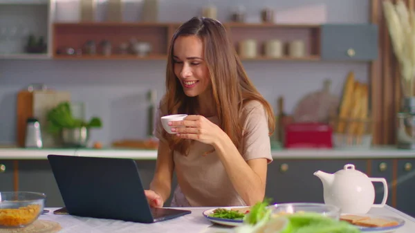 Nainen, jolla on video chat kannettavalla keittiössä. Tyttö juo kahvia kotona — kuvapankkivalokuva