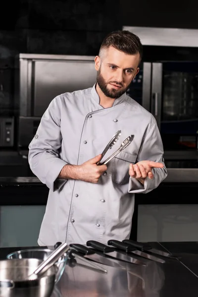 Szef kuchni trzyma szczypce w kuchni. mężczyzna kucharz pozowanie z metal tong. — Zdjęcie stockowe