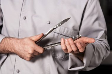 Chef prepare bbq at kitchen. Closeup male hands with metal tong. clipart
