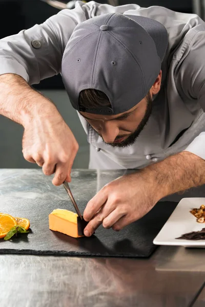 Chef macho cortar bolo no restaurante da cozinha. Chef homem decorar bolo de laranja . — Fotografia de Stock