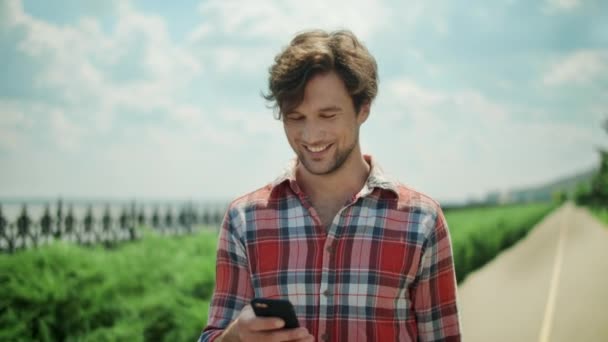 Lykkelig mand bruger smartphone udenfor. Smilende fyr går på vej i parken – Stock-video