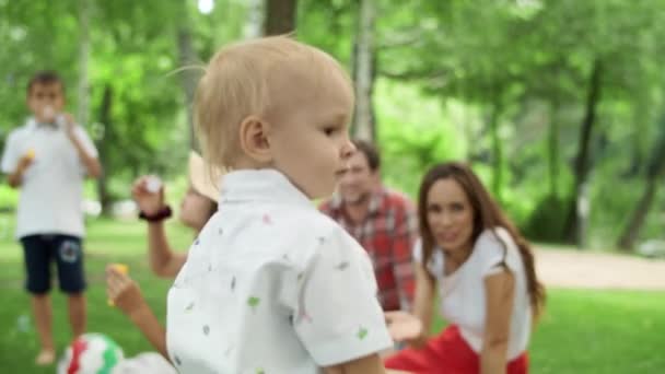 Тоддлер смотрит в камеру с семьей. Дети раздувают мыльные пузыри в лесу — стоковое видео