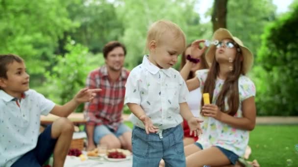 Liten småbarn poppar bubblor i parken med familj på bakgrunden — Stockvideo