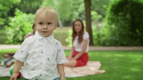 Rubia niño cubriendo la cara con las manos. Familia sentada en una manta en el parque . — Vídeos de Stock