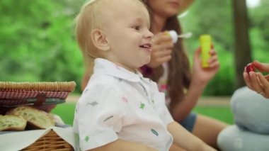 Anne ve kız kardeşiyle birlikte ormanda battaniyeye oturmuş gülen bir çocuk.