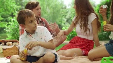 Ormandaki çocuklarla battaniyeye oturmuş. Kız ve erkek sabun köpüğü üflüyorlar.