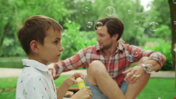 Los padres se relajan en la manta en el parque con los niños. Niños soplando burbujas de jabón — Vídeos de Stock