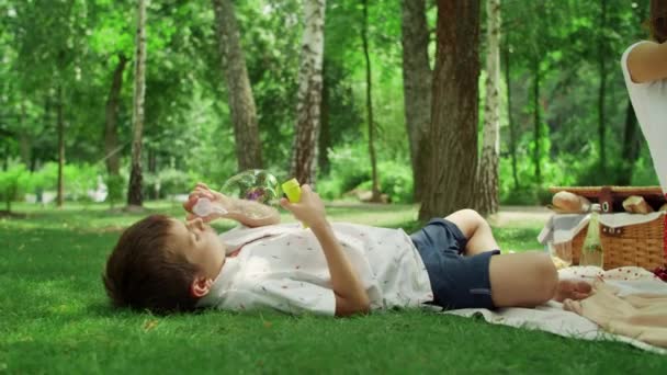 Imádnivaló fiú fekszik az erdő takaróján. gyerek fúj szappanbuborékok szabadban — Stock videók