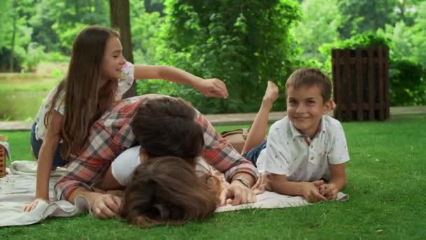 Семья лежит на одеяле в парке. Родители веселятся с детьми в лесу — стоковое видео