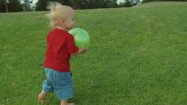 Imádnivaló kisbaba mezítláb megy a zöld mezőre. A fiú tartja a labdát a kezében — Stock videók