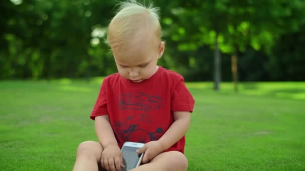 Niño sentado en el prado verde con teléfono. Lindo niño sosteniendo teléfono celular — Vídeos de Stock
