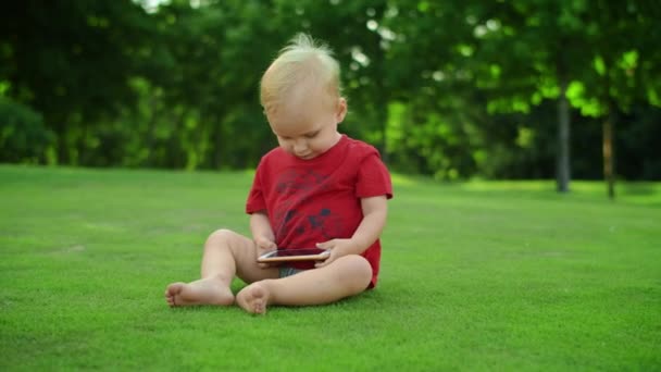 Очаровательный малыш держит в руках мобильник. Веселый мальчик сидит на зеленой траве — стоковое видео