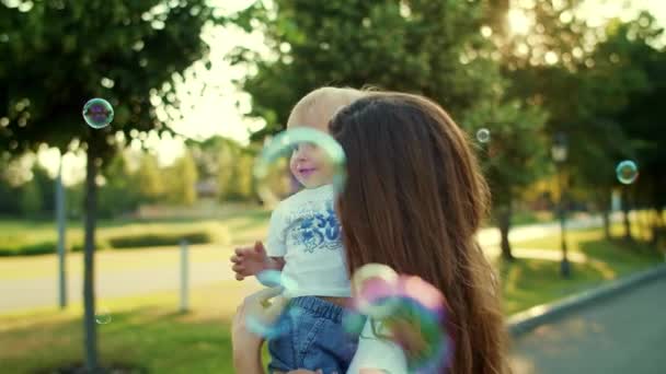 Мать держит сына на руках в парке. Женщина и ребенок играют с мыльными пузырями — стоковое видео