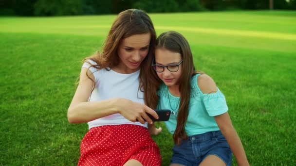 Женщина и девушка сидят на траве. Мать и дочь смотрят видео на смартфоне — стоковое видео