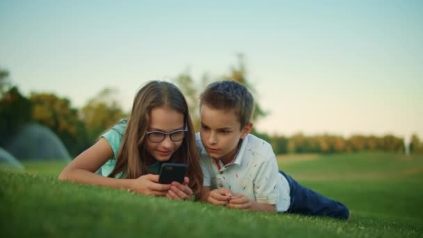 Παιδιά που χρησιμοποιούν smartphone στο πάρκο. Αδέρφια που βρίσκονται στο πράσινο γρασίδι στο λιβάδι — Αρχείο Βίντεο
