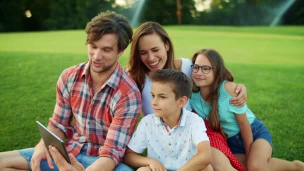 Familie sitzt auf Gras im Park. Eltern und Kinder telefonieren per Videotelefonie — Stockvideo