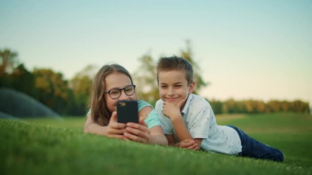Geschwister liegen im Gras auf dem Feld. Lachende Mädchen und Jungen mit Smartphone im Freien — Stockvideo