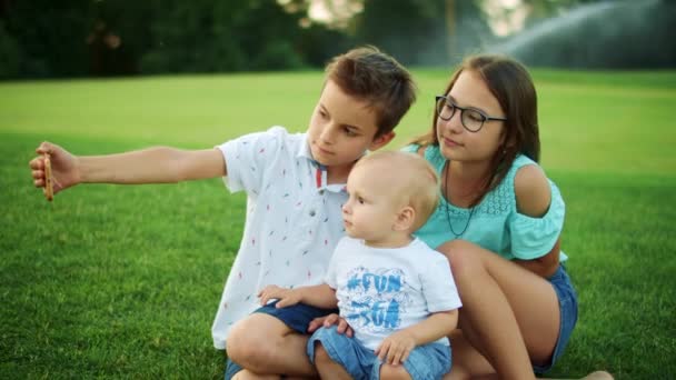 Usmívající se chlapci a dívka sedící na zelené louce. Děti berou selfie na smartphone — Stock video