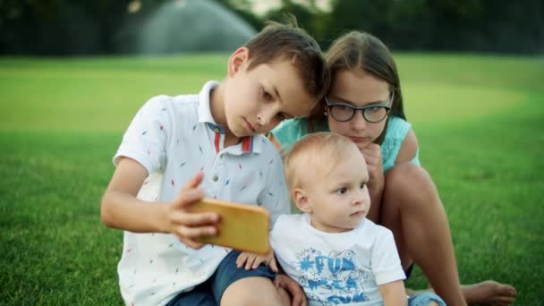Niños concentrados sentados en la hierba en el prado. Lindo chico tomando selfie en el teléfono celular — Vídeos de Stock