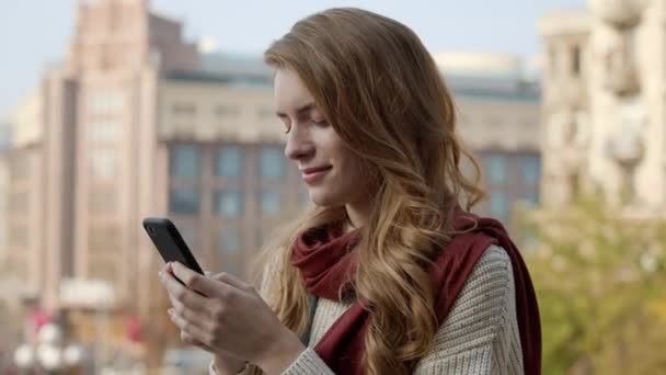 Kaunis nainen kirjoittaa tekstiviestejä puhelimitse ulkona. Söpö tyttö pitelee puhelinta kadulla. — kuvapankkivideo