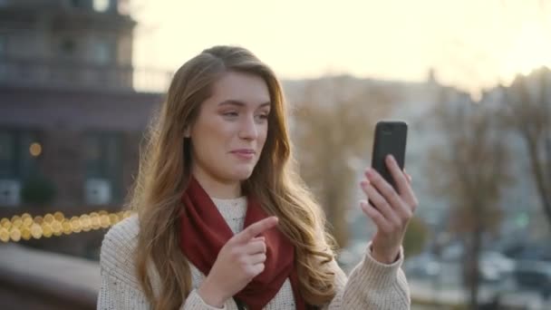 Улыбающаяся девушка с телефоном на улице. Женщина держит в руках мобильный телефон — стоковое видео