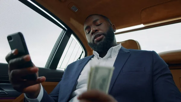 Nahaufnahme eines Mannes, der am Auto ein Video anruft. Afro-Mann schüttelt Geld auf Rücksitz — Stockfoto