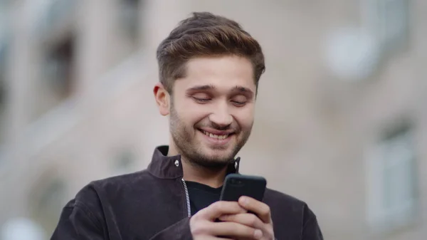 Um tipo sorridente a usar o telefone na rua. Homem feliz olhando tela do telefone fora . — Fotografia de Stock