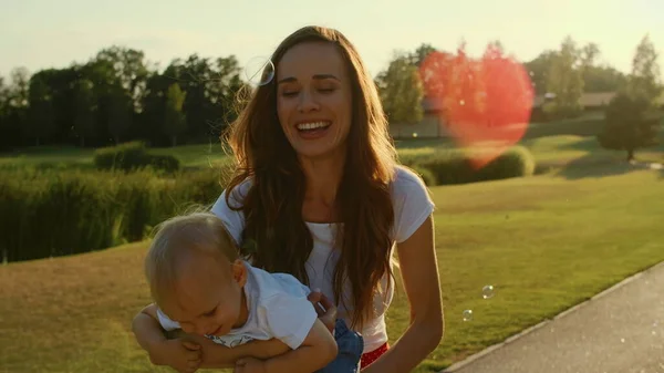 Moeder houdt kleine zoon op handen in park.Peuter op zoek zeepbellen buiten — Stockfoto